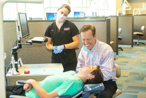 A boy in an orthodontist chair smiles at his orthodontist. An assistant with a mask is also smiling.