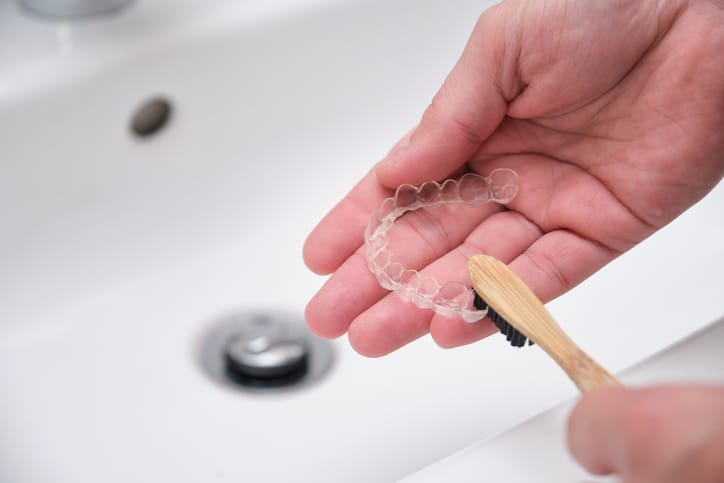 A hand cleaning a clear aligner with a toothbrush at the sink.