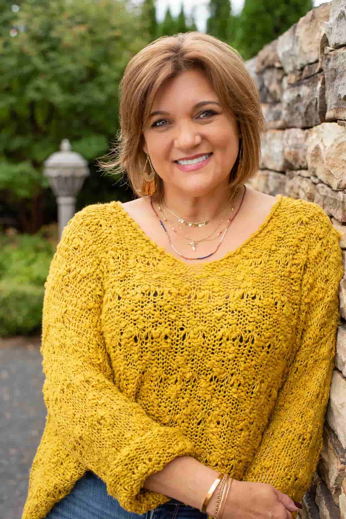 Woman with short brown hair and a yellow sweater, smiling outside next to a rock wall