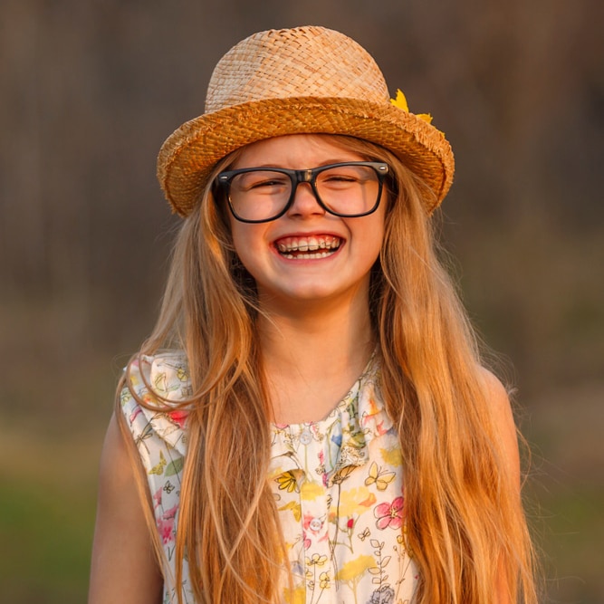 a child wearing glasses and a hat smiling and showing off her braces