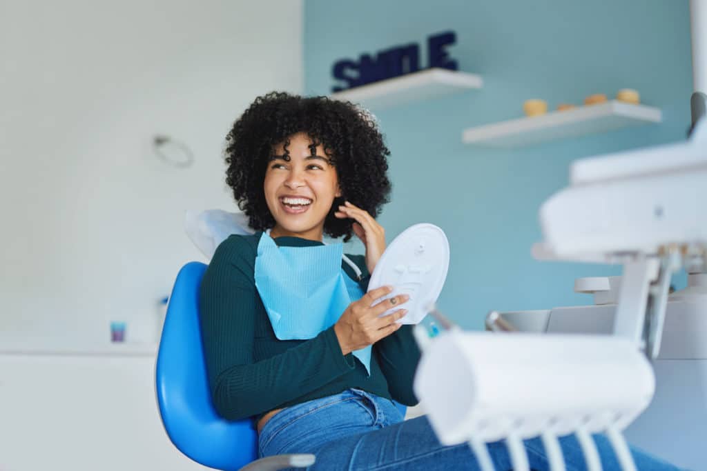 Girl with black curly hair sitting in a dental chair and smiling in a handheld mirror
