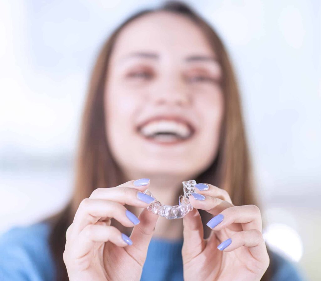 Smiling woman with lavender fingernails holding her invisalign aligner towards the camera