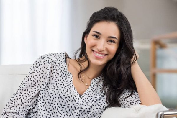 A teen girl with black hair and a black and white blouse sitting on a couch.