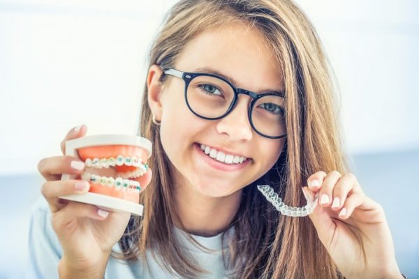 Girl with glasses holds a clear aligner in one hand, and a model of teeth with braces in the other.