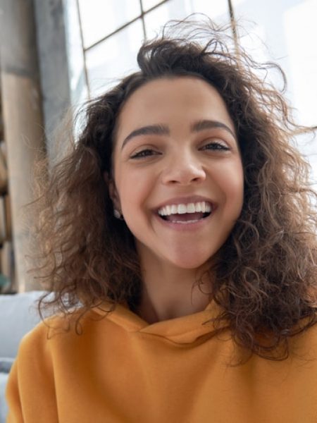 Young teen girl with curly hair and an orange sweatshirt. She is smiling and sitting in a window.