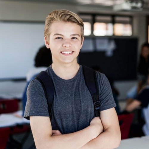 Portrait of smiling teenage boy standing against friends. Happy male blond student is with arms crossed in classroom. He is wearing casual clothing in school building.