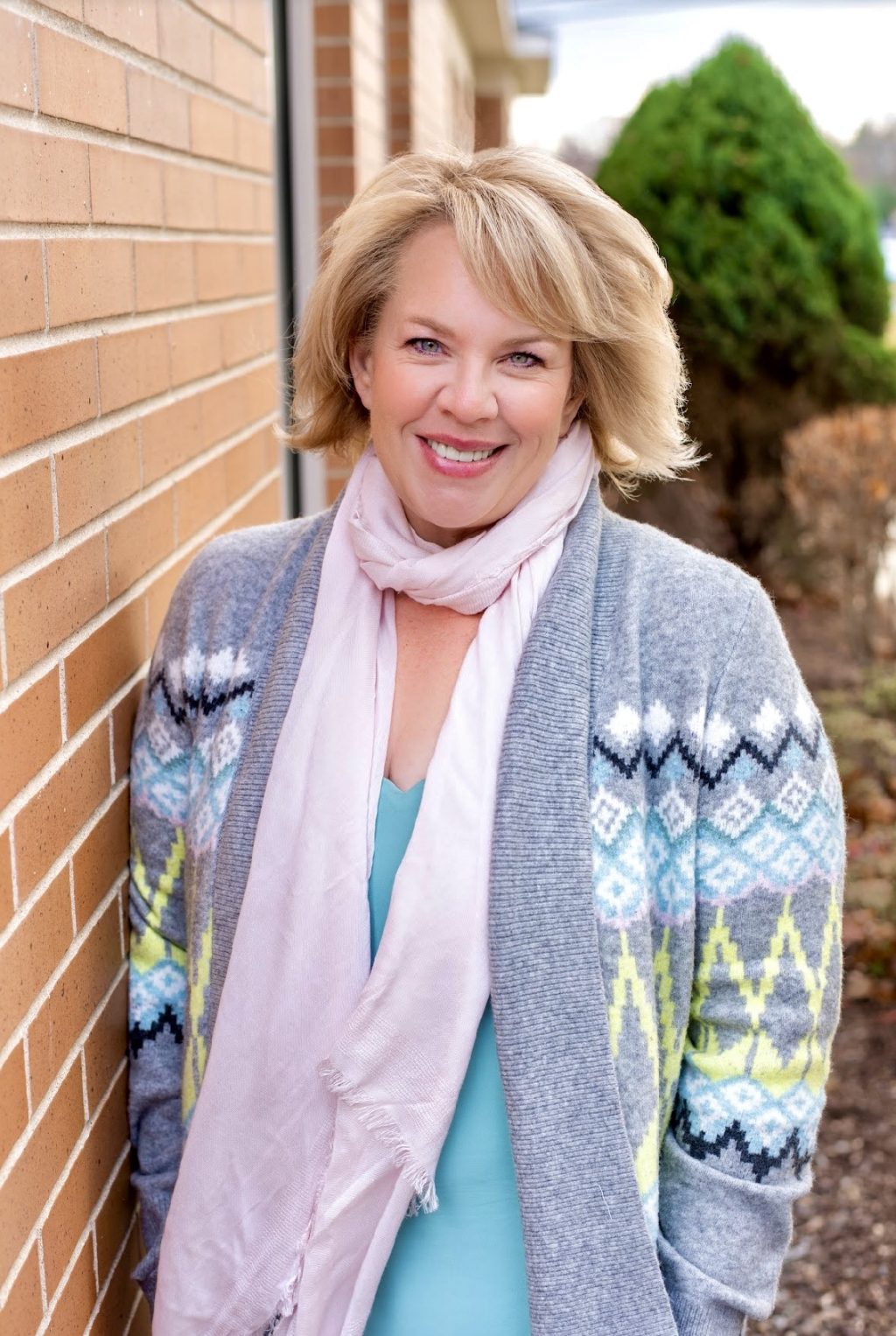 Woman with short blonde hair and a colorful sweater smiling next to a brick wall